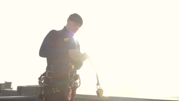 Vista da vicino di un arrampicatore professionista di sesso maschile in uniforme sul tetto che fissa chiusure e ganci con cinghie sul corpo per cavo e assicurazione. Una professione pericolosa per gli uomini coraggiosi ed estremi. — Video Stock