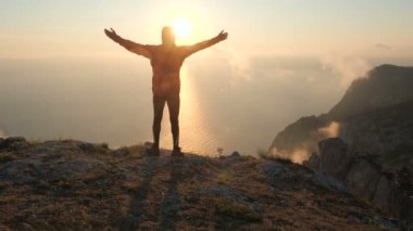 Dağın kenarında duran aktif bir adamın arkasından çekim yapmak ve gün batımı ve denizin muhteşem manzarasından kollarını yana açmak. Uçurumun kenarındaki turist erkek doğaya bakıyor..