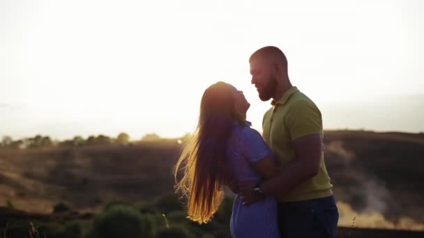 Cara barbudo abraços e beijos uma menina bonita inclinando no campo no pano de fundo do pôr do sol de verão. Férias românticas em conjunto na natureza de tarde de par jovem no amor em câmera lenta. Data fora da cidade . — Vídeo de Stock