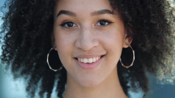 Hermosa joven africana confiada se ríe. Adolescente étnica feliz con ojos marrones, peinados afro rizados, pendientes perforados redondos, maquillaje ligero y sonrisas pecosas y muestra sus dientes blancos saludables. — Vídeo de stock