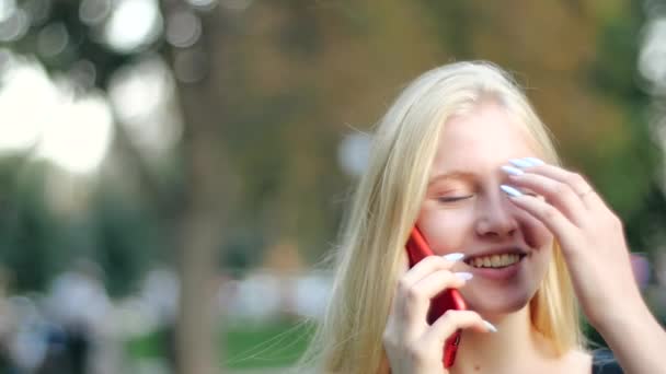 Uzun ipek saçlı, siyah şık elbiseli, güzel, güler yüzlü beyaz kız sıcak, güneşli bahar parkında kızıl akıllı telefonuyla saçlarını düzleştirirken mutlu bir şekilde konuşuyor. Cep telefonlu mutlu kız.. — Stok video