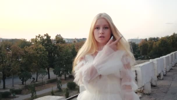 Acelerado cinemático primer plano disparo encantadora chica rubia linda con el pelo largo y sedoso y elegante vestido blanco caminando en la cubierta de observación contra el fondo de los árboles de primavera en el centro de Kharkov, Ucrania . — Vídeos de Stock