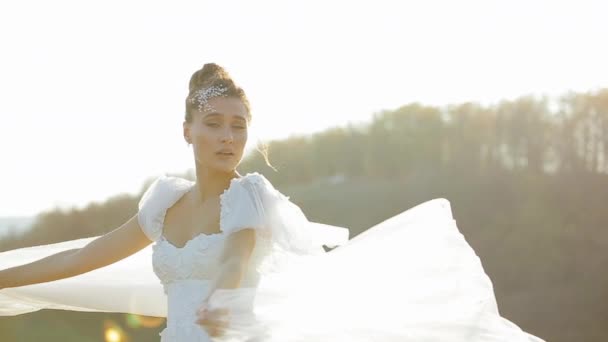 Sexy joven novia de apariencia europea se encuentra en un campo desierto y se refugia en un vestido de novia blanco largo y hermoso del viento sobre el fondo de la naturaleza borrosa en un día soleado de otoño . — Vídeo de stock