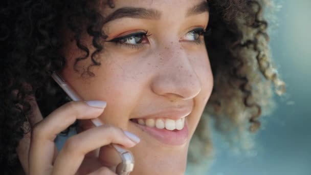 La mujer étnica americana joven habla por teléfono se alegra sonríe los dientes blancos, se acercan. Estudiante chica hace pedidos en línea en el teléfono inteligente fuera. Comunicación satisfecha con el cliente por teléfono — Vídeos de Stock