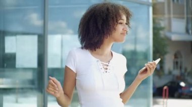 Etkin Afrikalı etnik kadın açık havada elinde akıllı telefonla müzikle dans ediyor. Eğlenceli duyguları olan mutlu kıvırcık kız komik aktiviteler yapmaktan hoşlanıyor, ağır çekimde. Modern yaşam tarzı kavramı