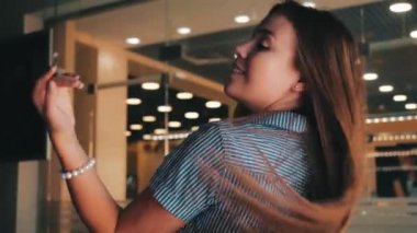 Çizgili elbiseli, güzel, gülümseyen genç bir kızın, aynadaki yansıyan ışıkların arka planına doğru uzun saçlarını yavaş çekimde salladığı yakın plan görüntüsü. Tatlı pozitif kız kamerayla flört ediyor..