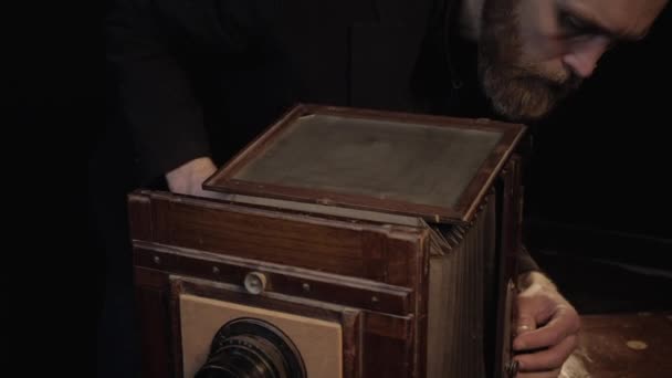 Homme barbu en veste noire ajuste soigneusement examine vieille caméra accordéon en bois rare sous la lumière de petits projecteurs en studio sombre isolé. Restauration, réparation de la technologie rétro antique obsolète — Video