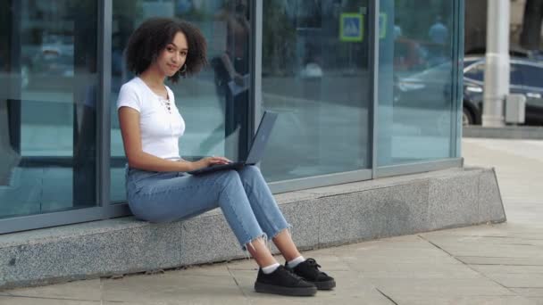 Pleasant young positive curly hair african american ethnic woman enjoying work at laptop smiling showing ok sign thumb up, gives an excellent mark of high quality sitting by reflective window outdoor — Stock Video