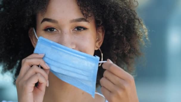 Hermosa sonriente joven afroamericana mujer poner máscara en la cara para proteger de la gripe coronavirus brote segunda ola de pandemia, mirando a la cámara al aire libre. Prevención de enfermedades en lugares públicos — Vídeos de Stock