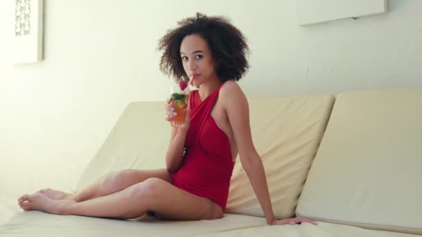 Jeune adulte frisée brune afro américaine dame en maillot de bain rouge assis sur la chaise de bureau salon à la maison hôtel complexe touristique ou lit de repos tenant et boire des fruits frais cocktail tropique regardant la caméra — Video