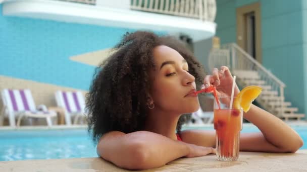 Expresión rizado bronceado afro chica en la piscina del hotel resort con cóctel de alcohol disfrutar de vacaciones de lujo en el extranjero estilo de vida exitoso. Cara hermosa coquetea mujer con jugo de frutas mirando a la cámara — Vídeos de Stock