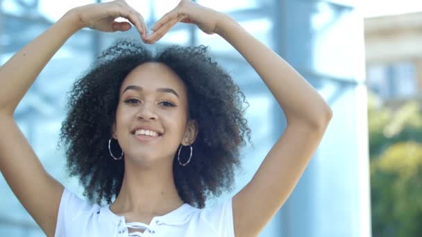 Sorridente giovane donna mostra segno in forma di grande cuore le sue mani. Concetto di carità, aiuto, amore, come speranza. Affascinante modello di ragazza afroamericana con trucco, capelli ricci posa con fiducia sulla strada — Video Stock