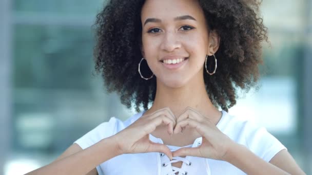 Bella giovane ragazza afro-americana sorride dentalmente, piega le mani nel segno del cuore di fronte a lei. Amichevole donna etnica sembra innamorato, concetto di come, fiducia, amicizia, carità, speranza. — Video Stock