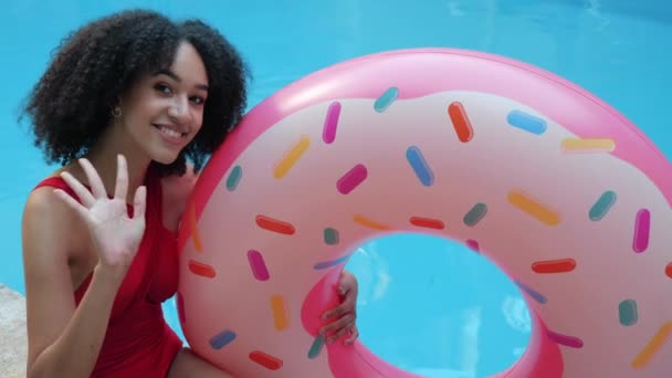 Счастливая американская этническая кудрявая женщина в красном купальнике сидит у бассейна, держа розовый надувной круг пончик улыбается, машет рукой, говорит привет удаленная связь глядя в камеру — стоковое видео