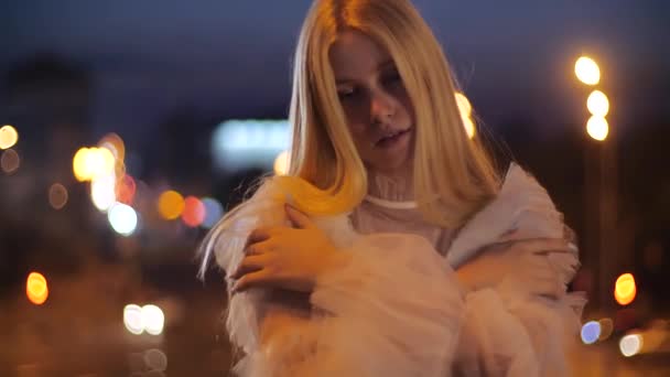 Schattig jong Kaukasisch meisje blond in licht witte jurk omhelst zichzelf met armen proberen warm te houden van de wind in de avond in het centrum van de schemering stad, close-up uitzicht. Mooi meisje poseert voor een foto. — Stockvideo