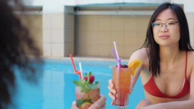Gözlüklü Asyalı etnik kadın ve kırmızı bikinili kıvırcık kadın havuz kenarında oturmuş meyveli kokteyl içerek neşe saçıyor ve gülümsüyor. İki kadın tesiste juce ile kadeh kaldırıyor.