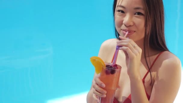 Portrait de femme asiatique joyeuse dans des lunettes assis au bord de la piscine en plein air boire cocktail sourire en regardant la caméra. coréen étudiant fille détente à l'étranger station test alcool boisson sur la plage, gros plan — Video