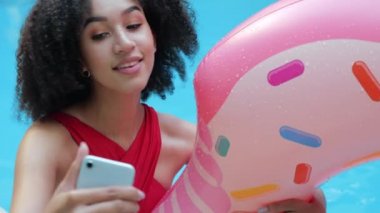 Havuzda dinlenen neşeli Afro-Amerikalı kadının portresi ıslak pembe şişirilebilir donut tutarak gülümsüyor cep telefonuyla internetteki sosyal ağlar için selfie ya da video yapmak için akıllı telefon kullanıyor.