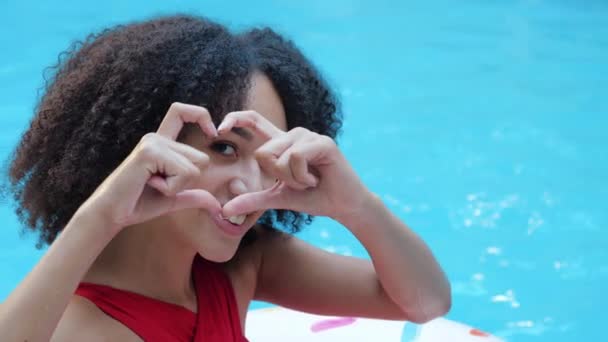 Jovem estudante Africano encaracolado mulher em repouso sentado perto da piscina sorrindo amigável olhando para a câmera fazendo dedos forma de coração dobrar as mãos em sinal de amor e enviar beijo de ar na câmera, close up retrato — Vídeo de Stock