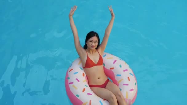 Sozinho jovem asiático fêmea em fato de banho vermelho nadando na piscina em círculo inflável donut rosa levanta as mãos sorri olhando para a câmera, tiroteio de cima. Etnia mulher relaxar na água no complexo turístico — Vídeo de Stock