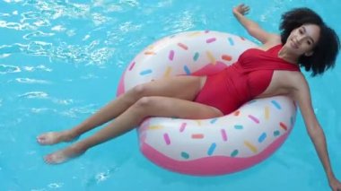 Bronzlaşmış kıvırcık kadın, pembe donutta yüzdükten sonra otel havuzunda dinleniyor. Kameraya gülümsüyor, yukarıdan ateş ediyor. Kırmızı mayo giyen Afro genç kız şişme halkalı mavi suyun keyfini çıkar.