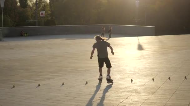 Ein junger langhaariger Rollschuhfahrer tanzt an einem schönen Abend im Stadtpark zwischen Zapfen. Freestyle Slalom Rollschuhlaufen zwischen Kegeln in Zeitlupe. — Stockvideo