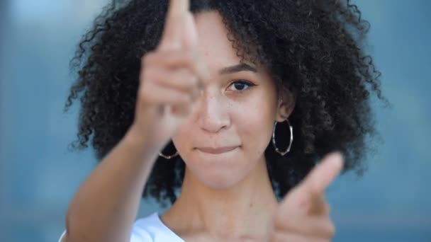 Encantadora chica adolescente afroamericana con estilo afro peinado hace pistolas de mano objetivo de golpe, finge disparar arma de los dedos sonrisa felizmente dentadura. Hermosa joven mujer coquetea divertidamente con confianza — Vídeo de stock