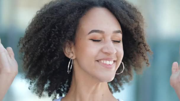 Mutlu heyecanlı Afrikalı Amerikalı kız kıvırcık saçlarıyla başını sallıyor, mutlu bir şekilde gülümsüyor ve zafer jesti olarak yumruklarını kaldırıyor. Genç bir kadın iyi haberlerden, samimi duygulardan ve başarıdan memnun olur.. — Stok video