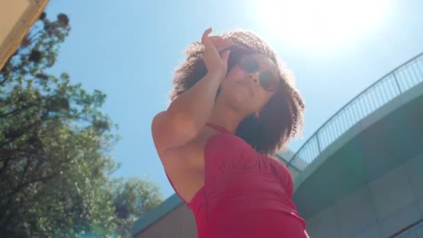 Modelo afro-americano bonito em maiô vermelho e óculos de pé ao ar livre perto do hotel ou restaurante no complexo turístico, tocando seu cabelo encaracolado, posando e flertando olhar para a câmera, bronzeando-se na vista de fundo do sol — Vídeo de Stock