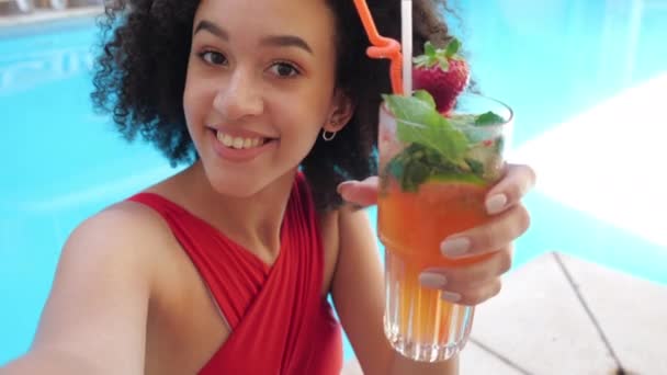 Zbliżenie szczęśliwej twarzy młoda blogerka Afro dziewczyna rozmawia z widzem lub przyjacielem online, siedzi w luksusowym basenie, pije koktajl owoców lodu z mięty i tworzenia zdjęć lub sieci wideo — Wideo stockowe