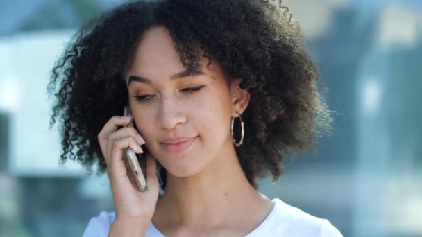 Linda menina adolescente afro-americana falando ao telefone, marcando uma consulta, esperando por amigos, amante, andando e relaxando. Mulher étnica percebe uma pessoa, feliz ondas e sorrisos, acena para ela . — Vídeo de Stock