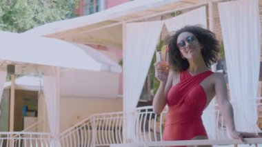 Havuz başındaki villa spa otelinin teras balkonunda duran, alkolsüz kokteyl içen yaz tatillerini kutlayan, selamlayan, kadeh kaldıran, kameraya gülümseyen modaya uygun mayo giyen çekici bir manken.