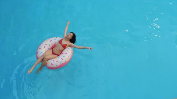 Draufsicht des schlanken jungen asiatischen Mädchens im roten Bikini-Badeanzug, das in einem aufblasbaren Rettungsring in rosa Donut liegt, die Hände hebt und im blauen Wasser schwimmt, sonniger Tag — Stockvideo