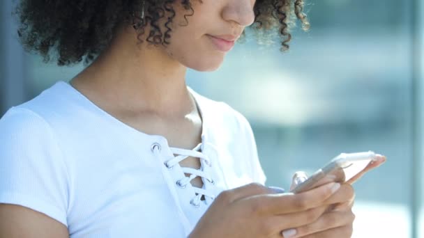 Gadis Afrika Amerika yang menarik tersenyum tajam, memegang smartphone di tangan, mengetik pesan di layar sentuh, berkomunikasi, mengirim pesan kepada teman-teman. Etnis muda wanita surfing internet, membuat ketertiban di toko — Stok Video