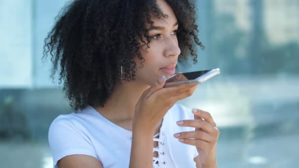 Hermosa chica adolescente afroamericana sosteniendo el teléfono móvil, hablando por altavoz y utilizando asistente virtual. Mujer étnica graba mensaje de voz a amigo, comunicación a distancia y pedidos en línea. — Vídeo de stock
