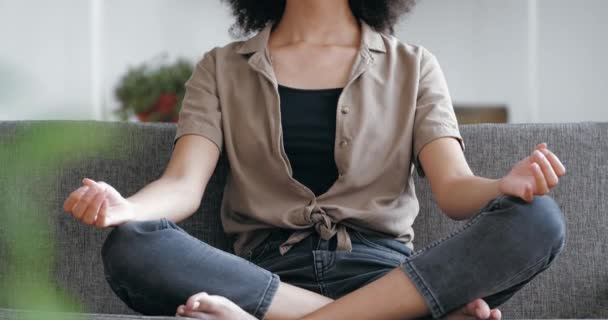 Afro donna americana si siede sul divano in Asana, medita, trova armonia. Donna razza mista in posizione di loto respirando profondamente, facendo esercizi di yoga, guadagnando equilibrio, alleviare lo stress, primo piano — Video Stock