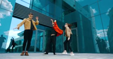 Üç genç dansçıdan oluşan bir grup aktif olarak birlikte hareket ediyorlar, ellerini sallıyorlar, modern koreografi binanın yakınındaki sokakta. İki erkek ve bir genç kız birlikte hareket ediyorlar, şehir manzarası.