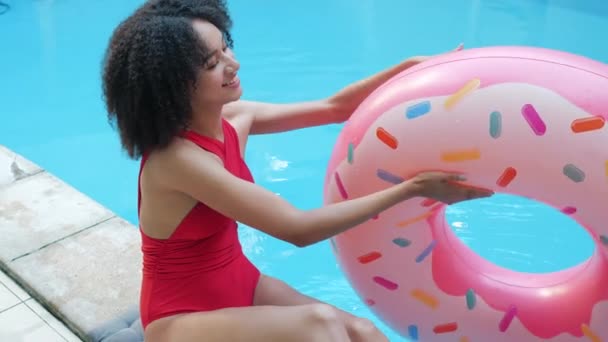 Giocoso allegro ridere ragazza afroamericana gioca con anello gonfiabile, getta gomma rosa ciambella in aria, rotei spin salvagente, indulge, in vacanza in piscina, guarda civettuola fotocamera — Video Stock