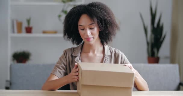 Emocionada sonriente mujer de raza mixta rizada recibe regalo, sostiene la caja de cartón abierta, mira dentro, sorprendido, disfruta de la entrega en línea rápida de la tienda en línea, descomprime el pedido sentado en casa en la habitación en la mesa — Vídeos de Stock