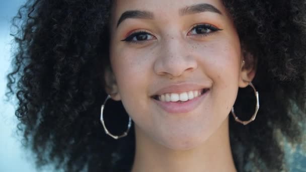 Bela jovem menina Africano confiante ri. Estudante étnico feliz com olhos castanhos, penteado afro encaracolado, brincos perfurados redondos, maquiagem leve e sardas sorrisos e mostra seus dentes brancos saudáveis — Vídeo de Stock