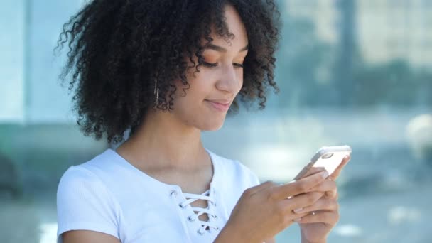 Lovely African American teen girl typing message to friend on phone, clicks on display, surfing Internet. Young woman looks at camera and smiles toothy, laughs at joke, makes online order in app. — Stock Video