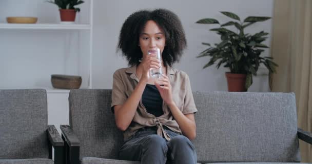 Joven mujer afroamericana sentada en el sofá en su casa en una habitación congestionada descansando relajada, saciando su sed con agua mineral fría y limpia en un día caluroso de verano, sintiendo un alivio refrescante — Vídeos de Stock