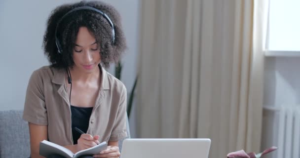 Afrikai diáklány otthon ül a kanapén, a szobából távolról tanul az e-tanulásról, elkülöníti a karanténidőt, fülhallgatót visel, hogy kommunikáljon a tanárral, jegyzetel, információt olvas. — Stock videók