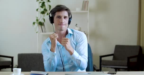 Lazy worker man dancing sitting in office at table, moving funny carelessly on break, resting from hard work. Freelance guy wears headphones and business shirt, listens music making wave with hands — Stock Video