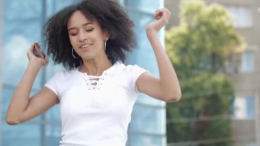 Neşeli genç Afro-Amerikan milenyum kadını yükseğe zıplamaktan, silah sallamaktan ve elinde akıllı telefonuyla havada dans etmekten, modern şehir binasının arka planına gülüp eğlenmekten hoşlanıyor..