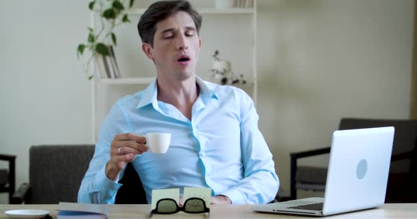 Молодой бизнесмен фрилансер сидит дома за столом в офисе, работает над ноутбуком, чувствует себя уставшим, растягивается, зевает, пьет чашку кофе, чтобы взбодриться, пытается сосредоточиться, рано утром концепция — стоковое видео
