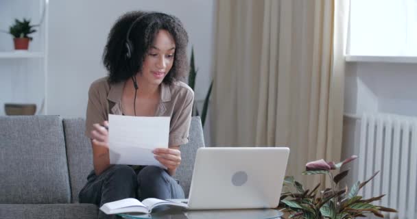 Молодая африканская женщина-предприниматель носит наушники, что делает конференц-звонок деловым, работая дома, сидя на диване. Далекая учительница из Америки ведет видеочат, общаясь с помощью веб-камеры на ноутбуке — стоковое видео