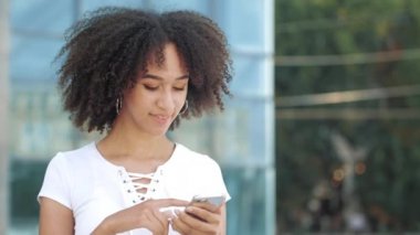 Gülümseyen Afro-Amerikan genç kadın akıllı telefona bakar, heyecan verici bir şekilde telefon ekranını işaret eder, beklenmedik bir sevinçle kendinden geçer, şehir çağdaş şehrinde heyecan verici duygular uyandırır..