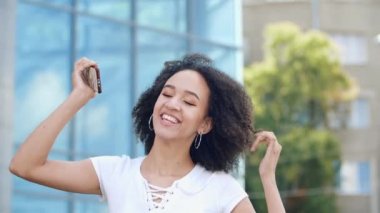 Mutlu, gülen, beyaz dişli, Afro-Amerikan mankeni yükseğe sıçrıyor ve elinde akıllı telefonuyla seviniyor, hareketten heyecan verici duygular yaşıyor, modern şehir sokaklarında inanılmaz duygular yaşıyor..