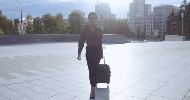 Vrolijk glimlachende Afro-Amerikaanse vrouw gaat straat naar het vliegveld met koffer, draagt paspoort en tickets in de hand. Gemengde race millenial dame aankomen na het reizen met bagage, zakenreis concept — Stockvideo
